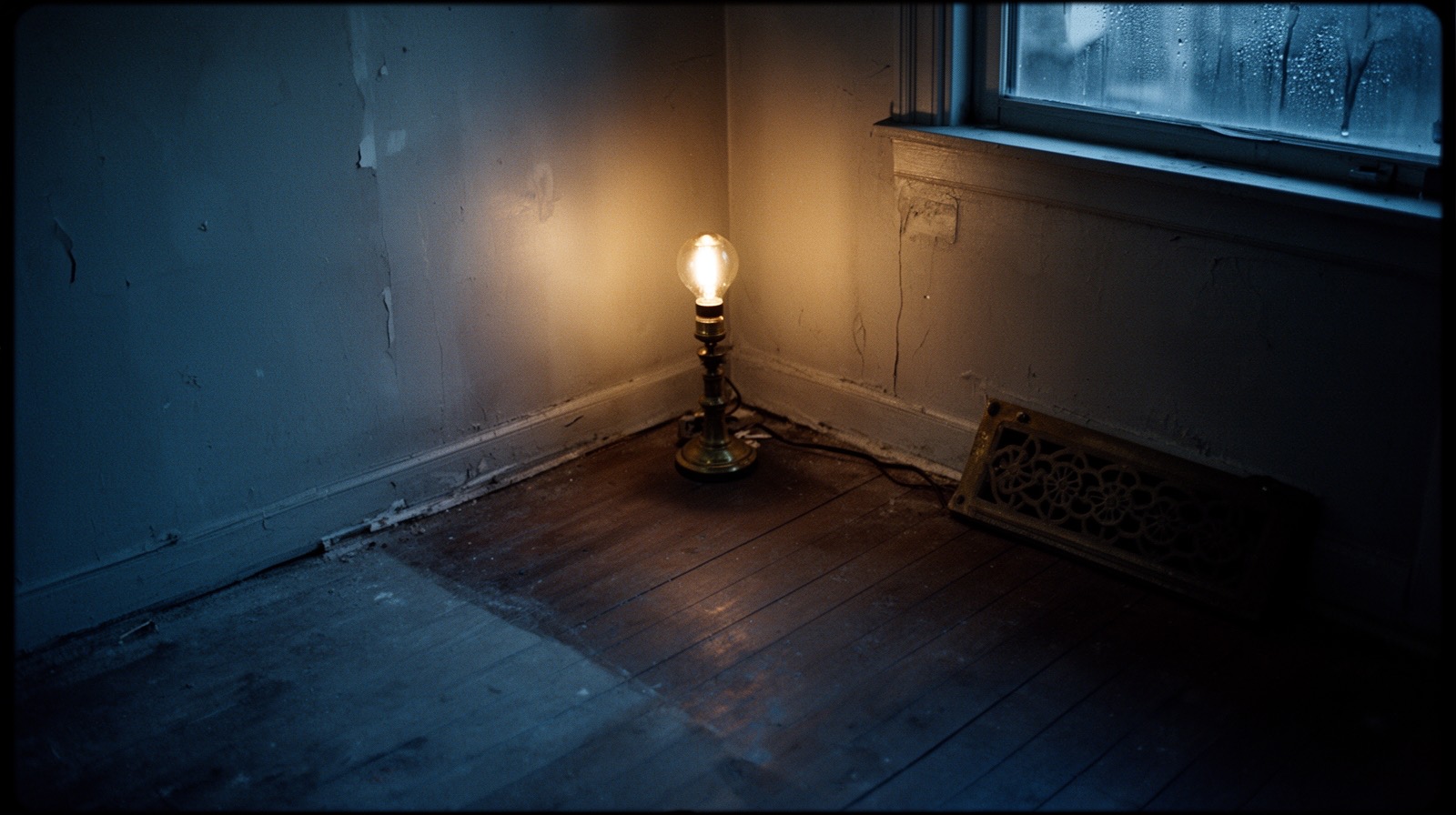 A dim, lamplit corner of an old house where one section of floor and wall sits in cooler, bluer light than the rest of the room.