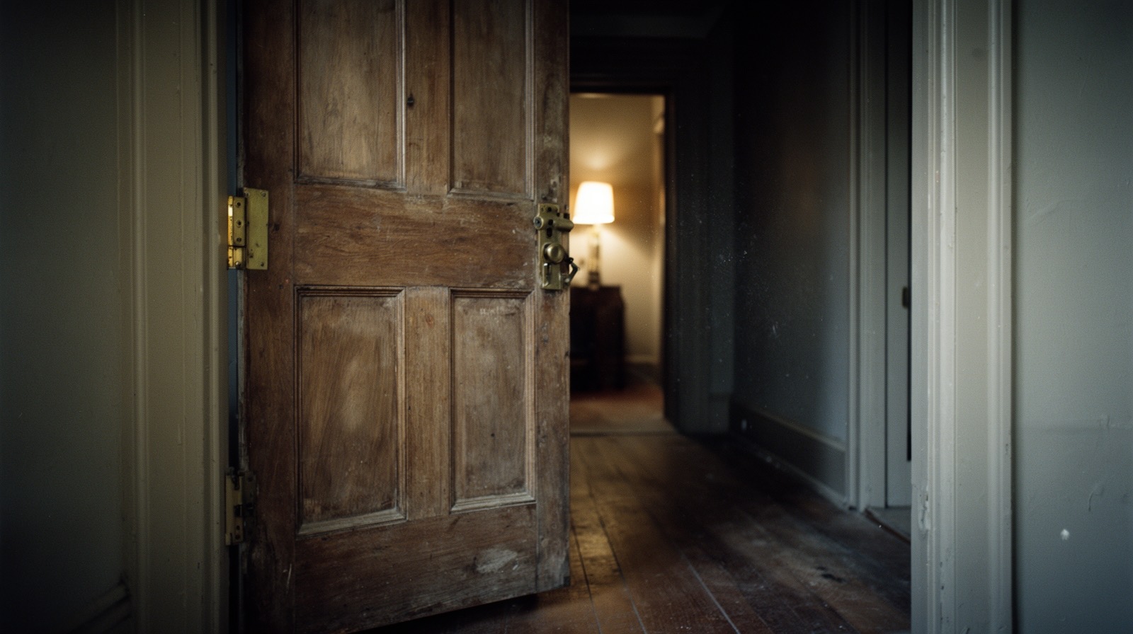 A wooden interior door standing ajar in a dim, lamplit hallway, caught mid-motion with no one nearby.
