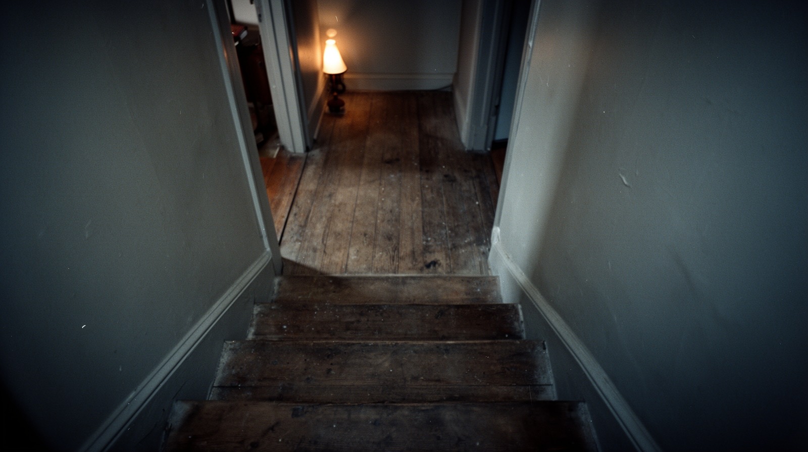A wood staircase leading to a dark upstairs hallway, lit only by a lamp's glow from an unseen room.