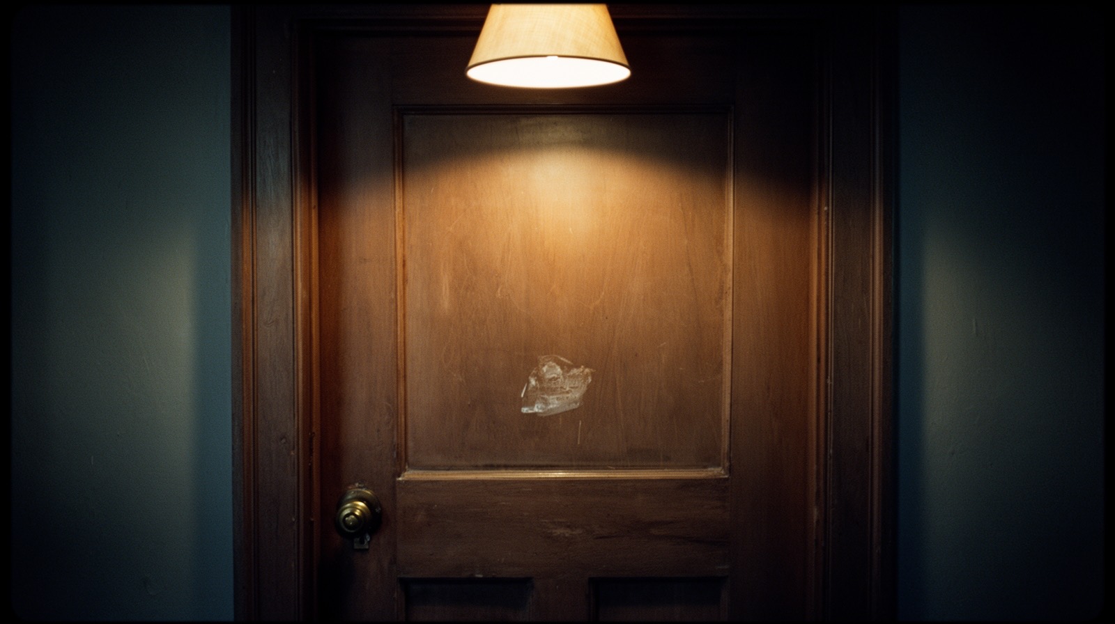 A paneled wooden interior door lit by a single warm lamp glow from the hallway, its upper panel faintly worn at knuckle height.