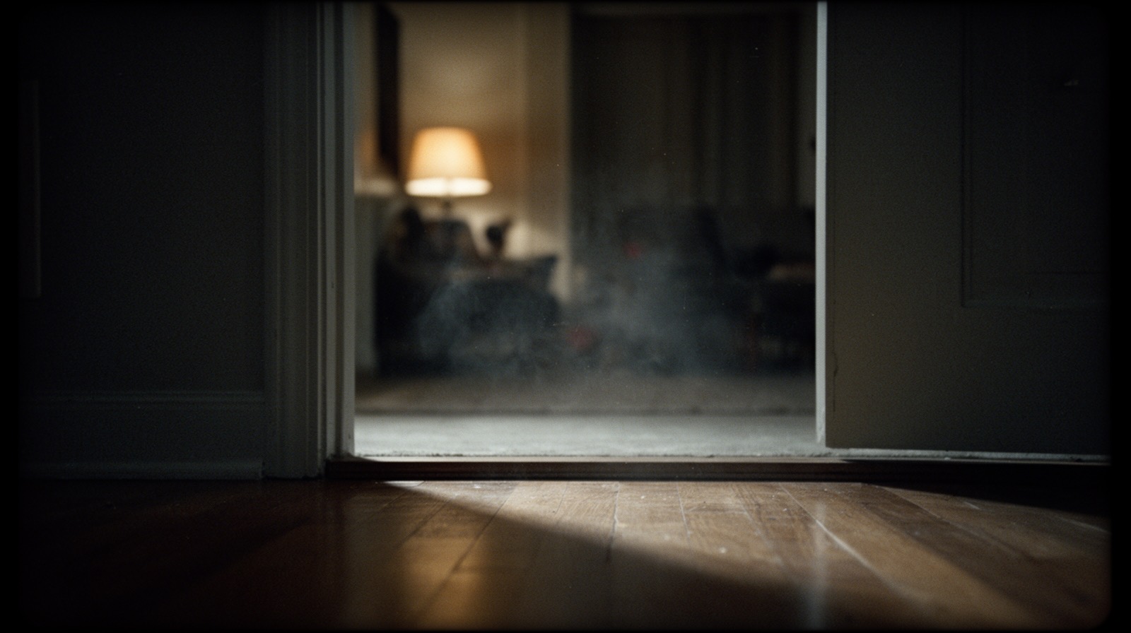 An empty doorway seen from floor level in a dim, lamplit room, with a strip of bare floor in sharp focus in the foreground.